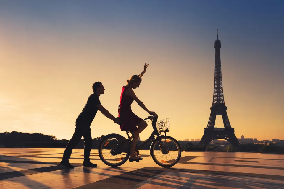 Silhouette of man holding on to back of bicycle ridden by woman with Eiffel Tower in background.